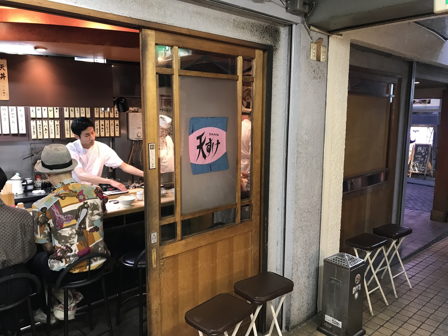 木枠の引き戸に「Tempura 天すけ」の青いのれんが貼られ、奥のカウンターで白衣の店主が客に天ぷらを提供している外観と店内の様子
