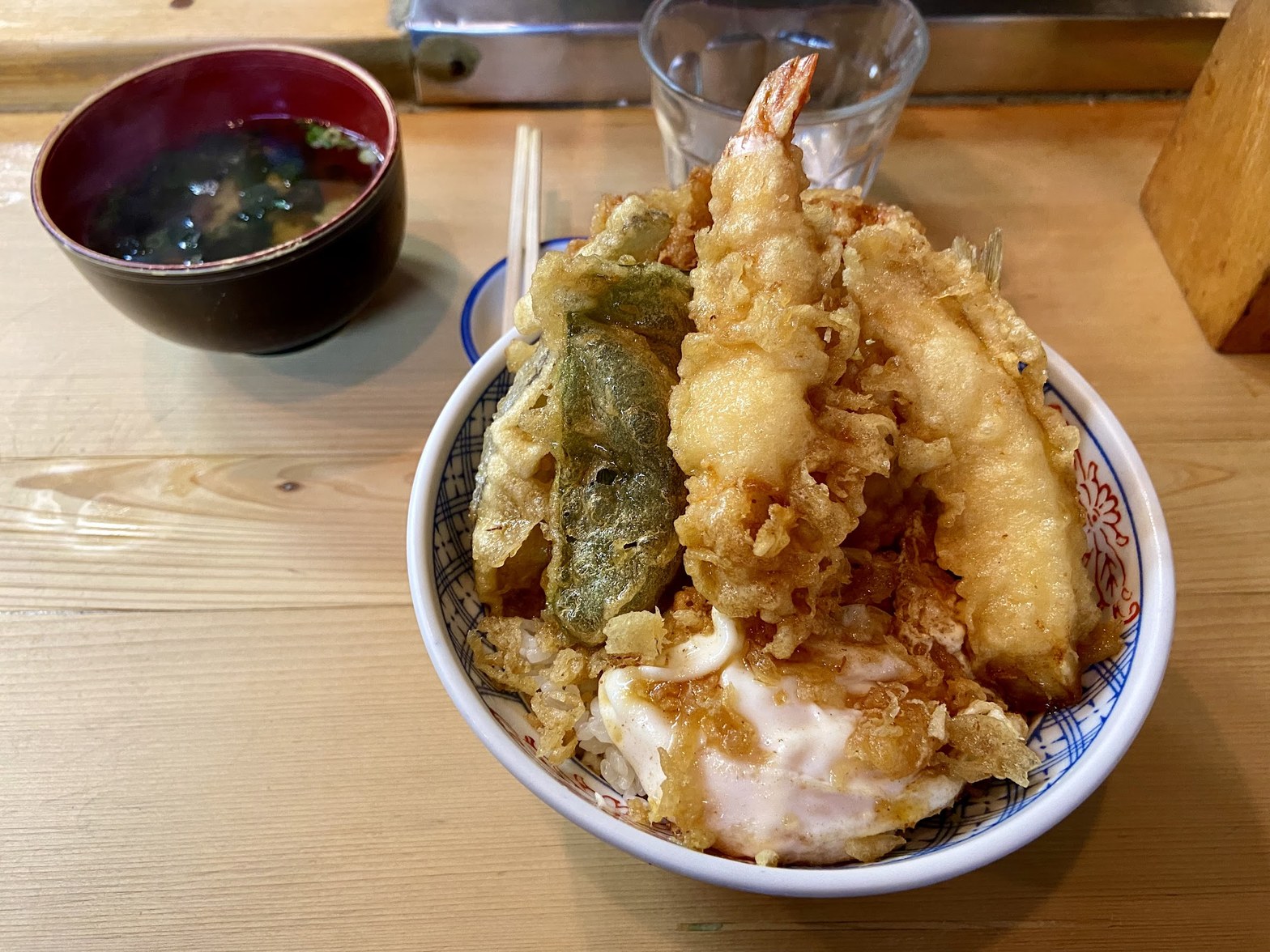 青柄の丼鉢に海老天・ピーマン天・かき揚げが盛られ、タレがかかった玉子天丼と赤い椀の味噌汁が木製カウンターに並ぶ
