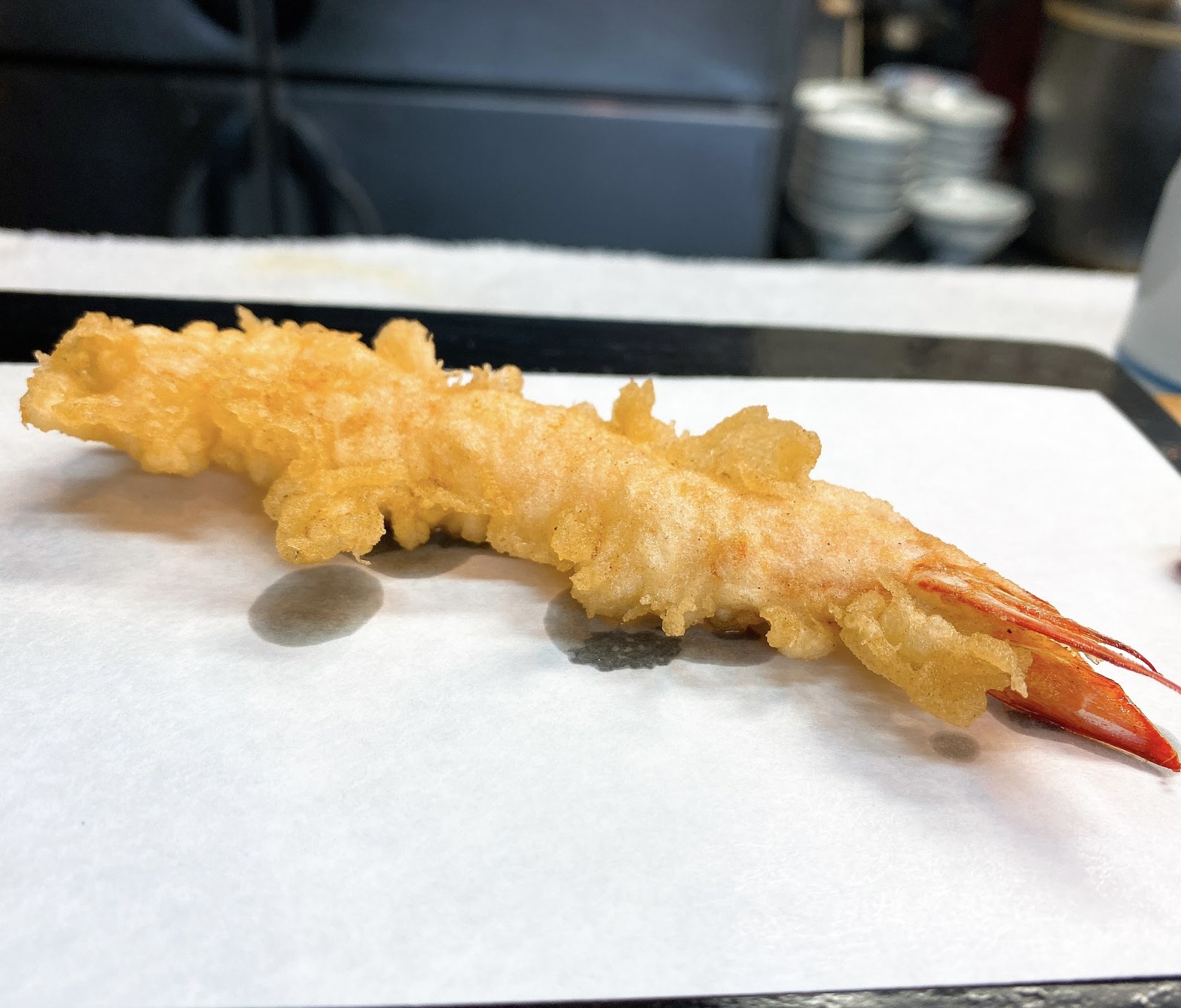 白い紙の上に置かれた揚げたての海老天一本、衣がサクサクに膨らみ尾の赤が際立つ